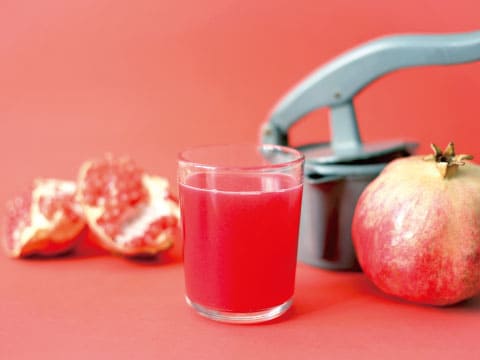 A glass of red pomegranate juice beside a hand-press juicer, a whole pomegranate, and pomegranate seed clusters on a red background