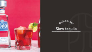 A deep red Slow Tequila cocktail in a rocks glass with mint and a lime wheel, set beside a tequila bottle against a pink background
