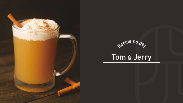 A hot Tom and Jerry cocktail in a glass mug topped with whipped cream and a cinnamon stick, garnished with a dusting of spice