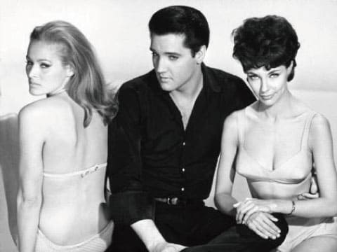 Vintage black-and-white publicity photo of a man in a dark shirt seated between two women in swimsuits