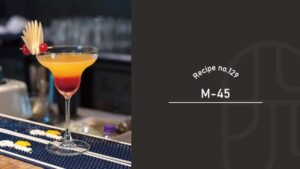 M-45 cocktail with an orange-to-red gradient in a margarita-style glass, garnished with an apple fan and red cherries on a bar mat