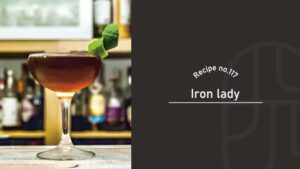 Iron Lady cocktail in a coupe glass with a deep amber-red color and a green citrus peel garnish on a bar counter with liquor bottles behind