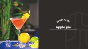 Apple Pie cocktail in a cut-glass martini glass, a soft coral-pink drink garnished with a twisted yellow citrus peel, beside an orange, a lime and ice cubes on a blue mat — Recipe no.152