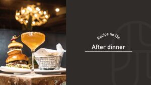 After Dinner cocktail with a golden amber color in a rimmed coupe glass, served beside a stacked burger and a basket of fries under a warm, glowing chandelier