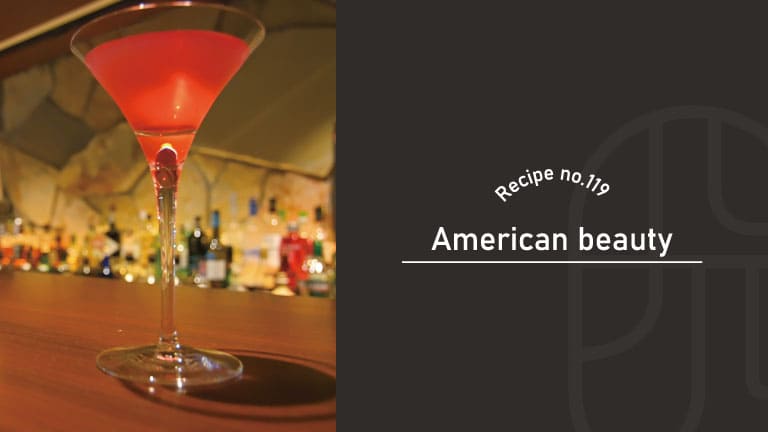 American Beauty cocktail with a vivid rose-red color in a tall-stemmed cocktail glass on a wooden bar counter with liquor bottles behind