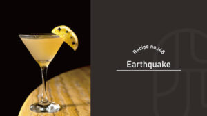Earthquake cocktail in a martini glass, a pale golden drink garnished with a lemon wedge studded with cloves, on a polished wooden table against a black background — Recipe no.148