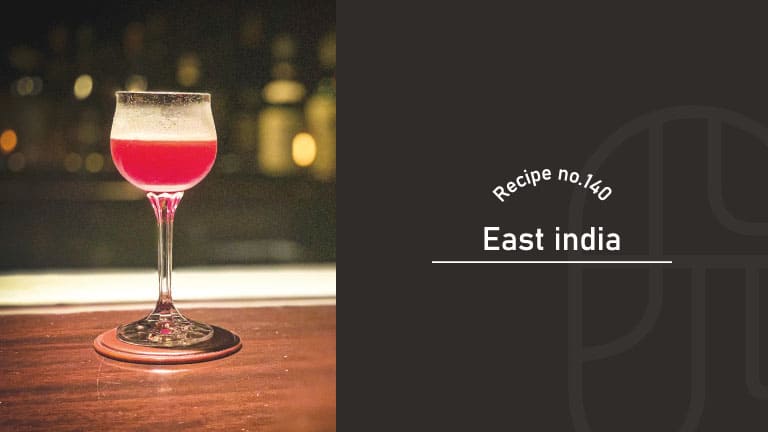 East India cocktail in a stemmed tulip glass, a deep ruby-red drink with a thin layer of foam, set on a leather coaster at a dimly lit bar — Recipe no.140