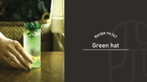 Green Hat cocktail in a tall highball glass, a pale green sparkling drink over ice with a lemon slice and a fresh mint sprig, held by a hand on a wooden bar table — Recipe no.147