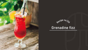Grenadine Fizz cocktail with a vivid ruby-red color in a hurricane glass, garnished with an orange slice and a black straw on a rustic wooden table outdoors