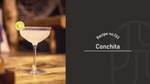 Conchita cocktail with a pale, cloudy color in a frosted-rim coupe glass, garnished with a lime wheel on a wooden bar counter