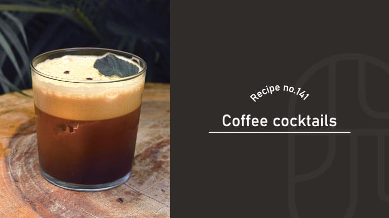 Coffee cocktail in a clear tumbler with a thick creamy foam top, garnished with a dark green leaf and coffee beans, on a rustic wooden board — Recipe no.141