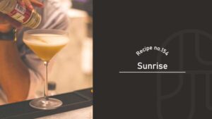 Sunrise cocktail with a frothy golden top in a stemmed coupe glass, as a bartender sprinkles a spice garnish over it at the bar, Recipe No.154