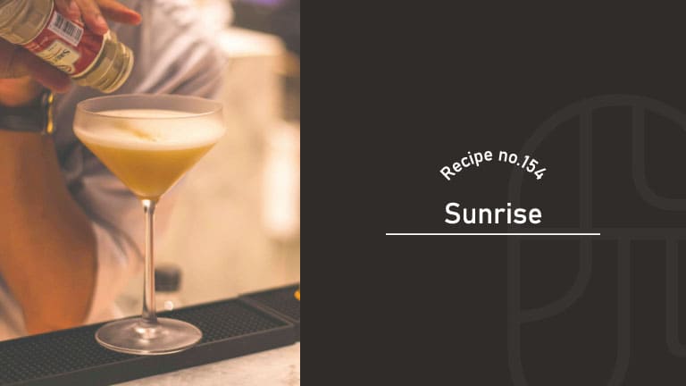 Sunrise cocktail with a frothy golden top in a stemmed coupe glass, as a bartender sprinkles a spice garnish over it at the bar, Recipe No.154