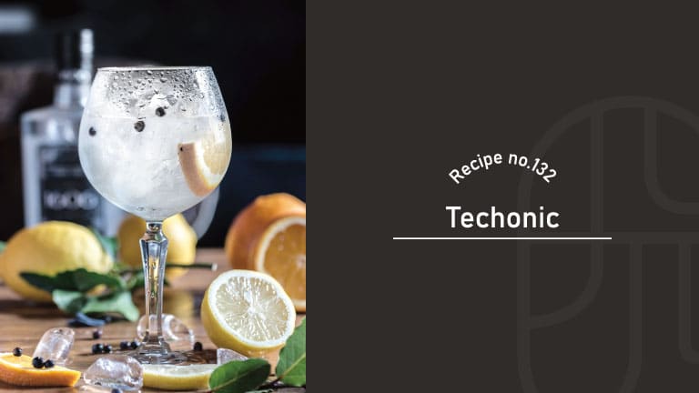 Techonic, a tequila and tonic cocktail, served sparkling over ice in a balloon glass with a lemon slice, surrounded by fresh lemons, oranges and green leaves on a wooden table
