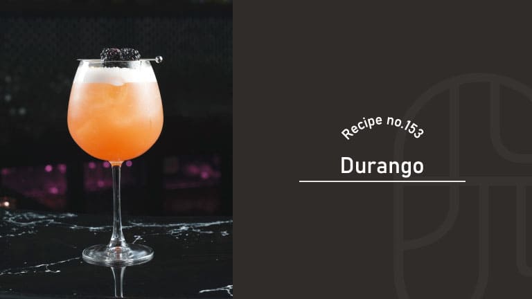 Durango cocktail in a balloon wine glass, a peach-orange drink with a thin layer of white foam, garnished with blackberries on a pick, on a dark marble bar — Recipe no.153