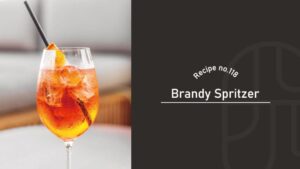 Brandy Spritzer cocktail in a wine glass with ice, an orange slice and a black straw, glowing amber-orange on a bright café table