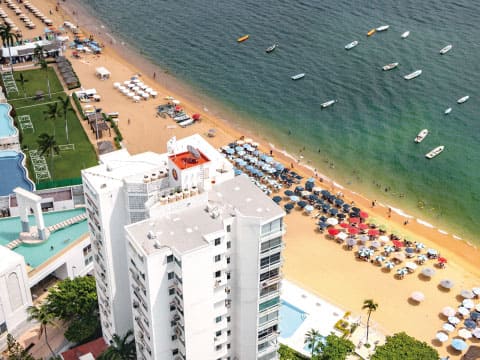 Aerial view of a sunny beach in Acapulco, Mexico, with white hotel towers, colorful beach umbrellas and boats floating in the calm blue bay
